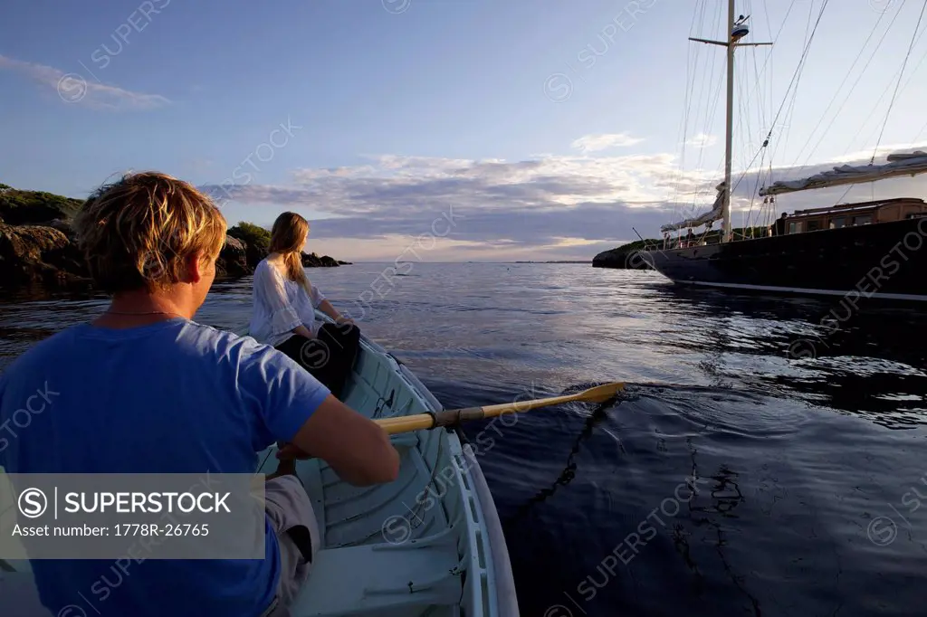 A young man and girl row in a Peapod dinghy at sunset toward a classic yacht moored along the coast of Maine.