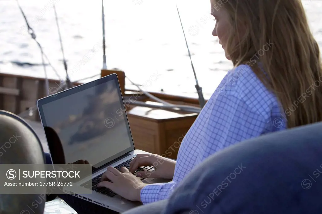 A teen girl uses a laptop computer on board a sailing yacht as the sky at sunset is reflecting on the screen.