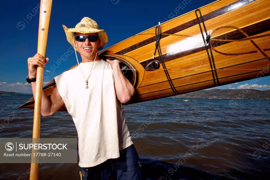 A man wears a straw hat as he poses for a portrait with a wooden kayak and paddle that he made while standing on the shore of Bear Lake, Utah.