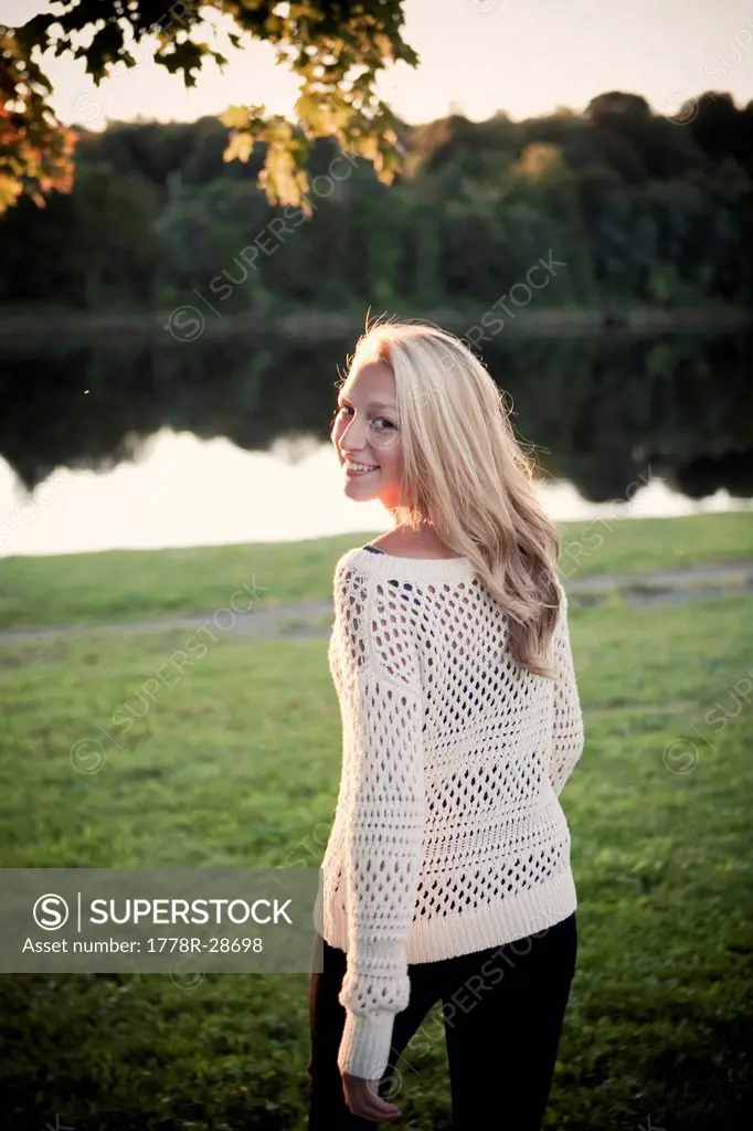 A young woman smiles and looks over her shoulder on the grassy bank of a river in the late afternoon sun.