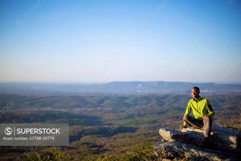 A young man sits on the ledge of a rock overlook on top of Mount Cheaha.