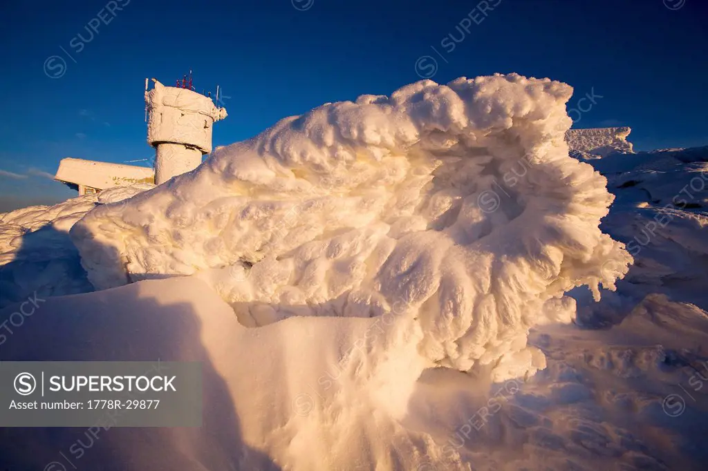 Large, jagged snow and ice formation on the summit of Mt. Washington with the Mount Washington Observatory in the background.