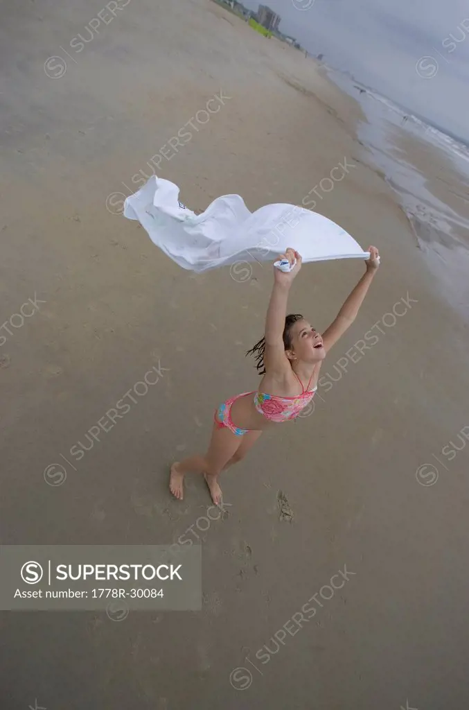 A young girl frolics on the beach with her towel and the blowing wind.