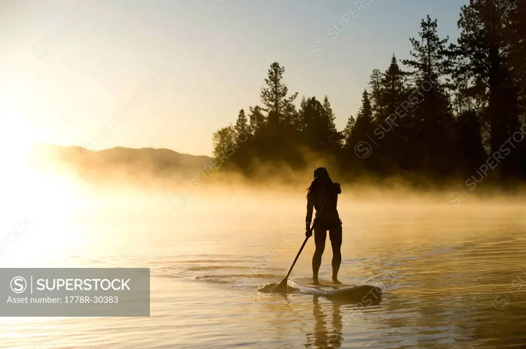 A woman, is silhouetted Stand Up Paddleboarding (SUP) at sunrise in the mist in Lake Tahoe, CA.