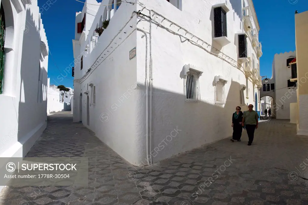 A man and woman walk down a street in the coastal town of Asilah, Morocco.