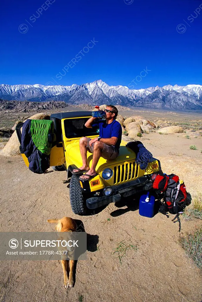 Man sitting on a 4_wheel drive vehicle in the desert with his dog taking a drink from a water bottle.