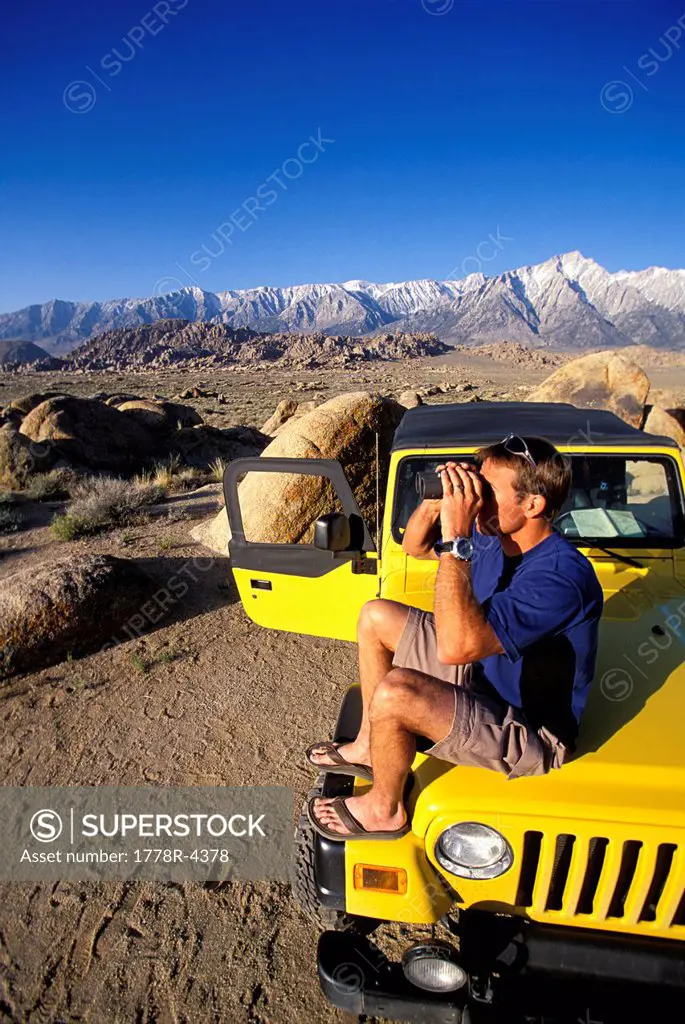 Man sitting on a 4_wheel drive vehicle in the desert next to mountains with binoculars.