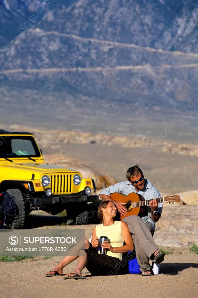 Man and woman sitting next to a 4_wheel drive vehicle in the desert playing a guitar.