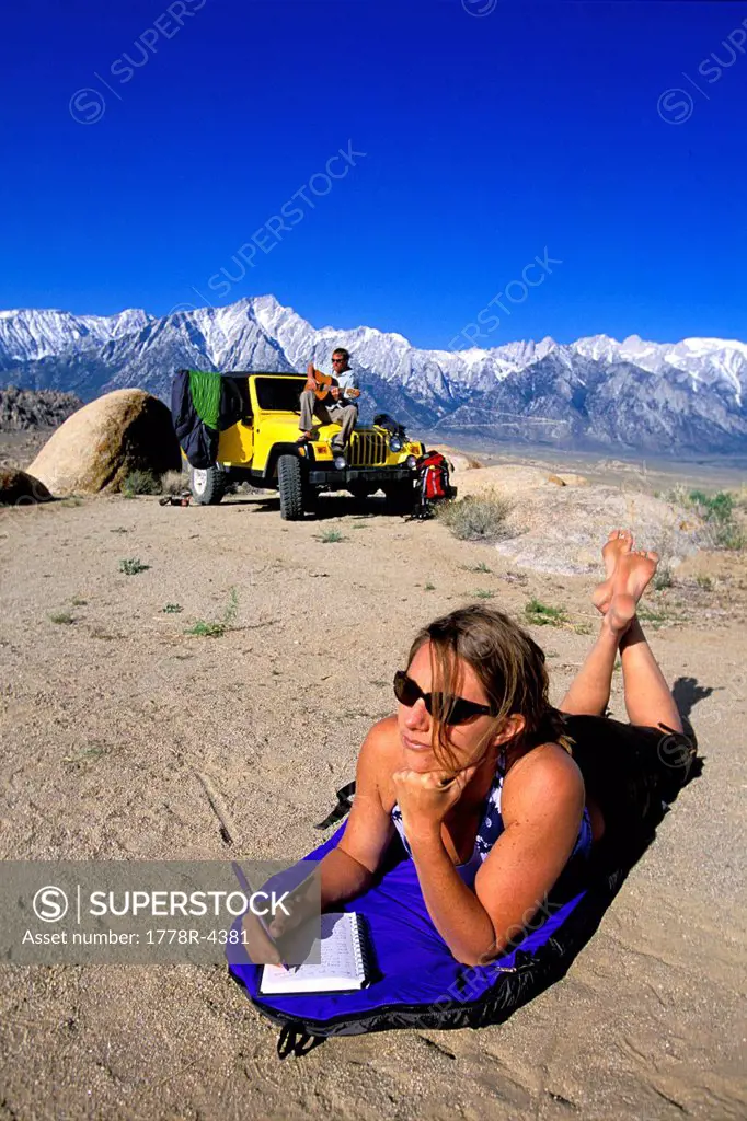 Woman laying on a sleeping pad in the desert with a man, 4_wheel drive vehicle and mountains in background.