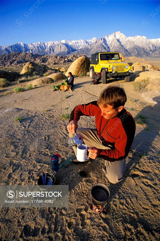 Man making coffee on a camp stove in the desert with a woman,4_wheel vehicle and mountains in the background.