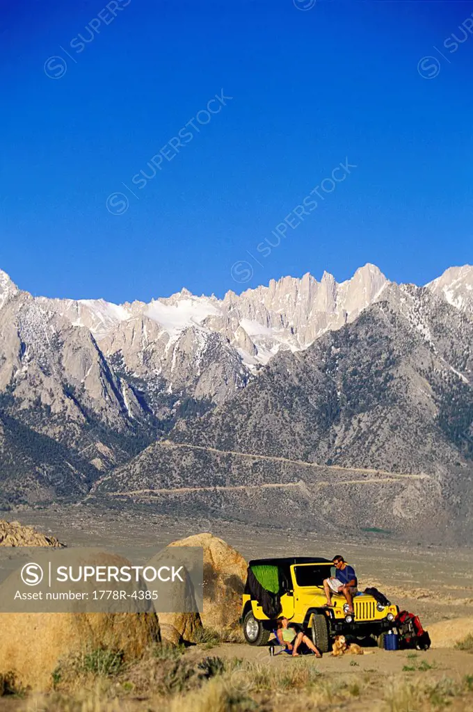 Man and woman hanging out next to a 4_wheel drive vehicle in the desert with mountains in the background.