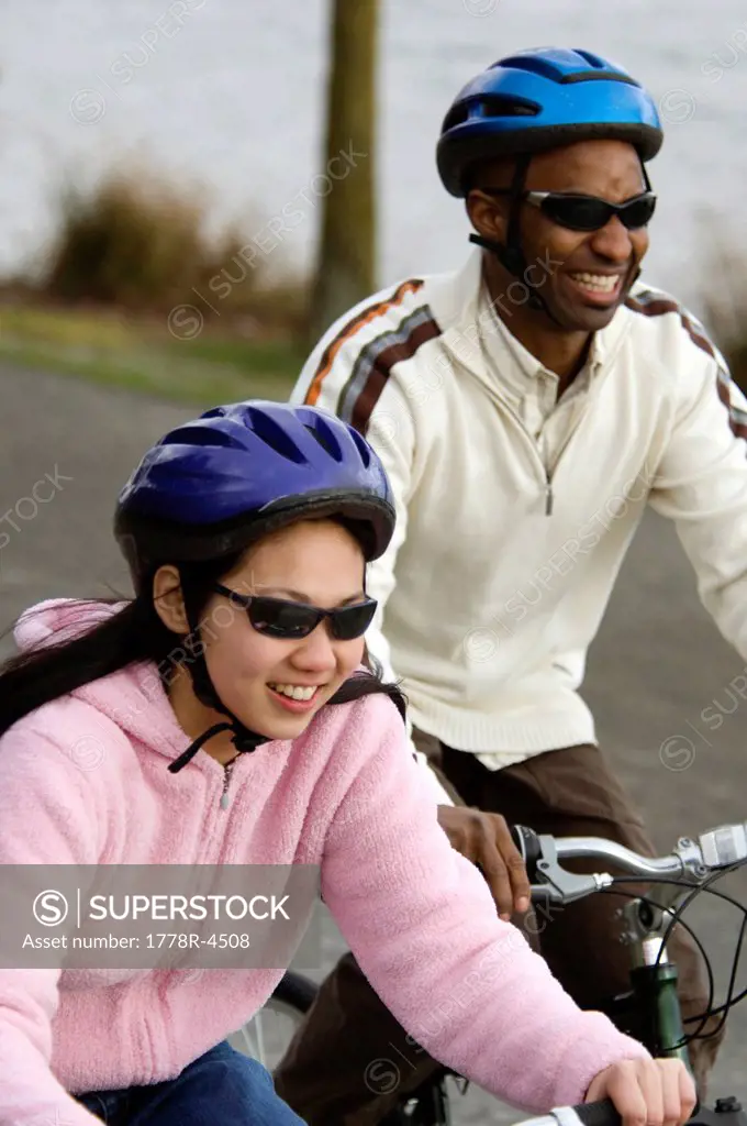 A young couple in helmets ride bikes on a path near the ocean.