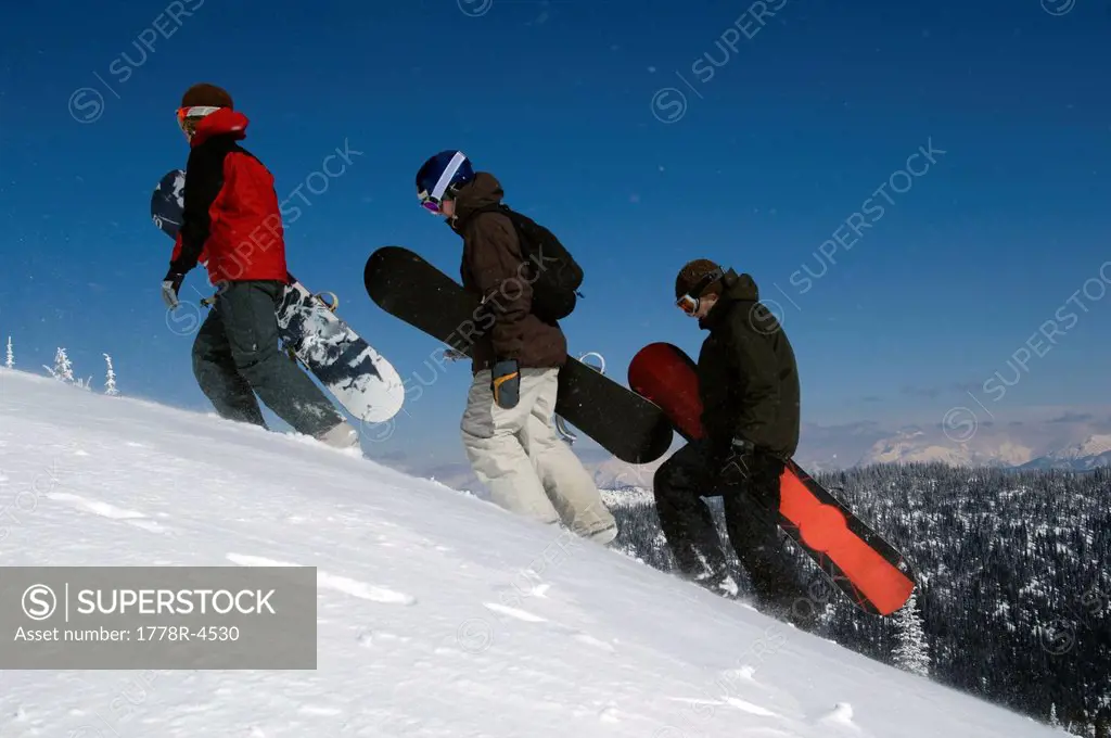 Three young men walk up a snowy mountain with their snowboards in the backcountry.