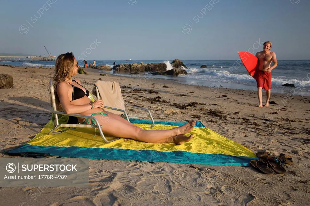 A woman sits in a chair on the beach while a man walks with his surfboard.