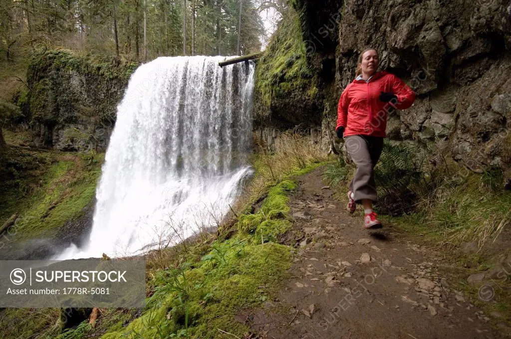 A woman trail running next to a waterfall in Silver Falls State Park, Oregon, USA.