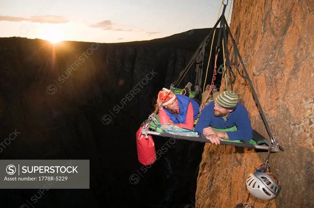 A man and woman on a portaledge at sunset while rock climbing a vertical face in Gunnison, Colorado.
