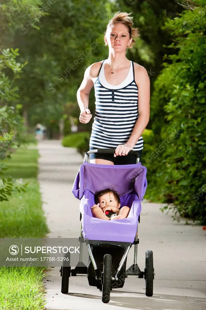 An edgy_looking, athletic young woman enjoys a run on a suburban sidewalk with a baby in a jogging stroller.