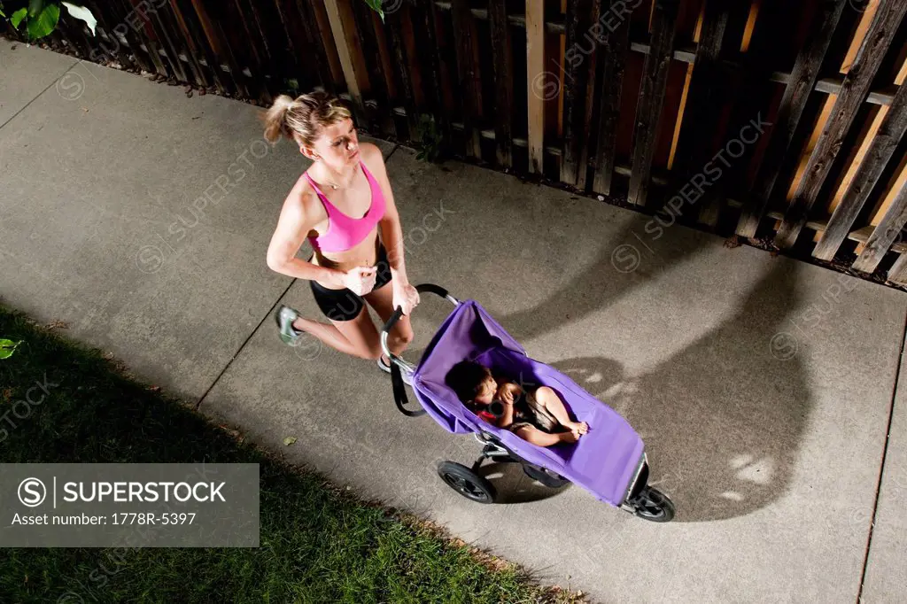 An edgy_looking, athletic young woman in a sports bra enjoys a run on a suburban sidewalk with a baby in a jogging stroller.
