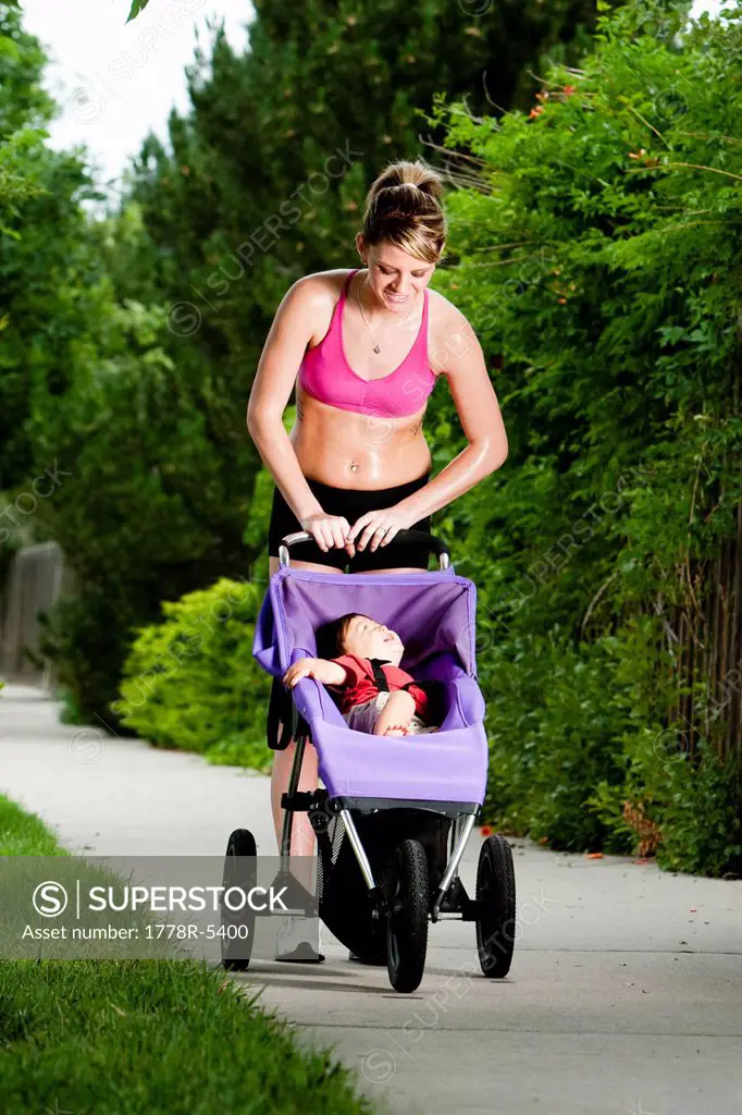 An edgy_looking, athletic young woman looks down at a smiling baby in his jogging stroller during a run on a suburban sidewalk.
