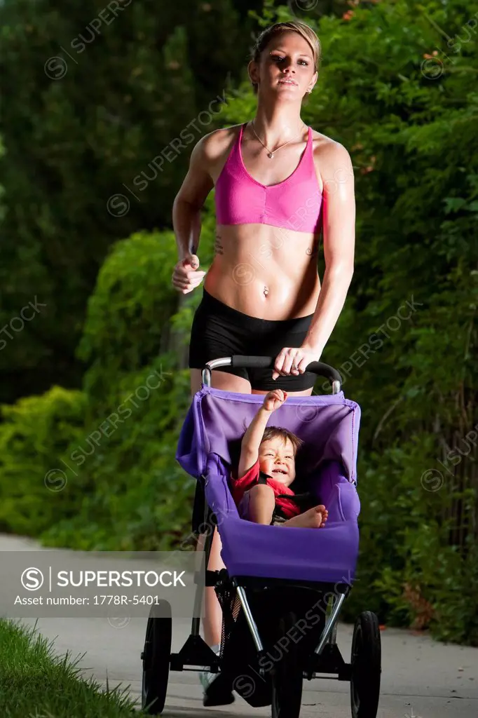 A baby raises his fist and smiles in his jogging stroller while being pushed by an edgy_looking, athletic young woman during a run on a suburban sidew...