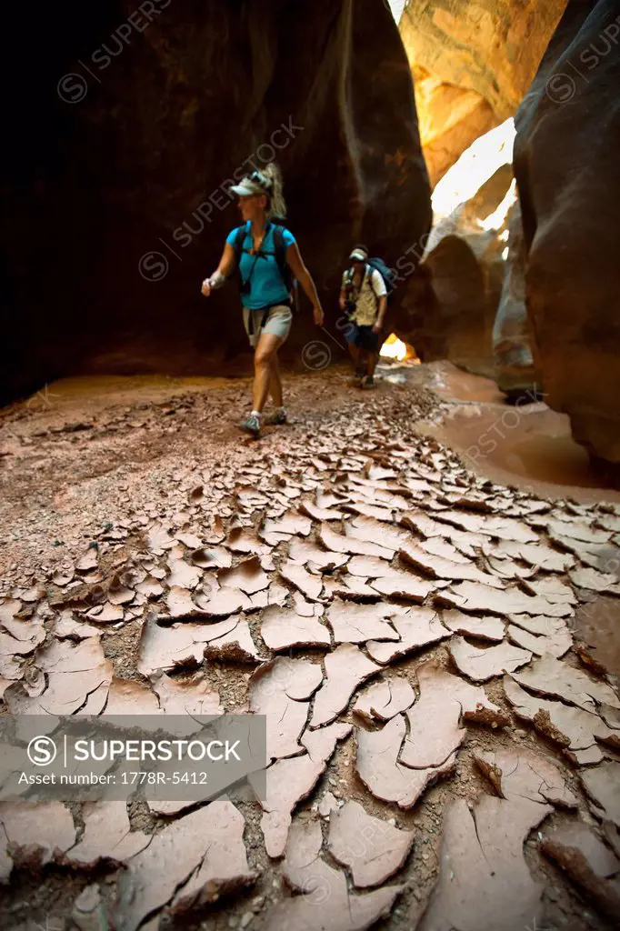 A man and woman hiking through a narrow canyon past dried and cracked mud in Utah.