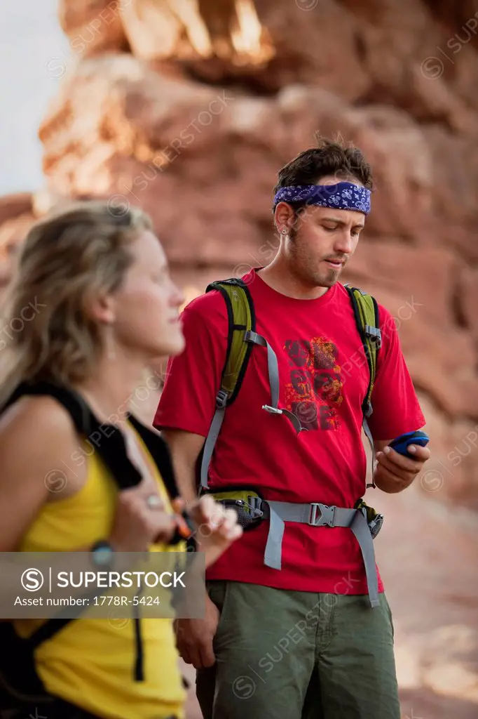 A man with backpack on looks at his GPS while an out of focus woman in the foreground looks on.