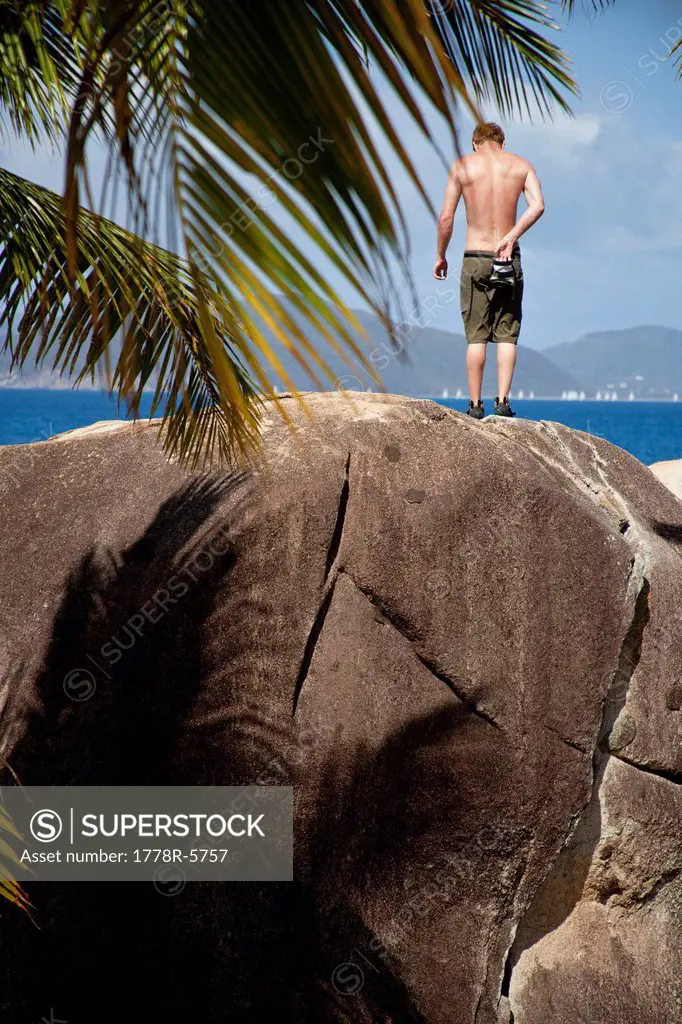 A male climber stands at the top of a problem on the beach surrounded by palm trees in the British Virgin Islands.