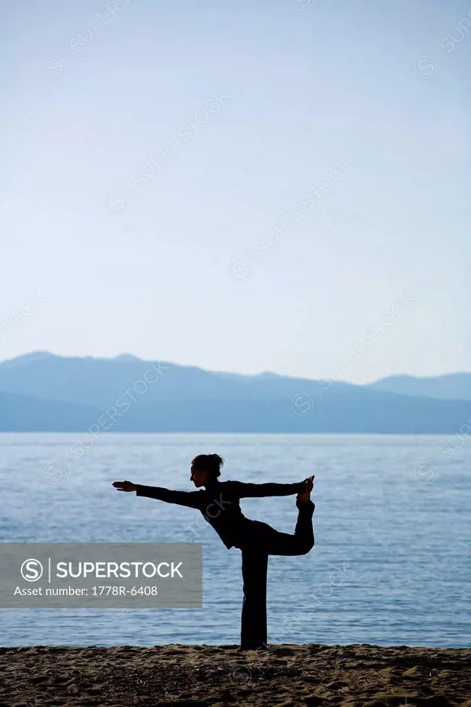 Silhouette of a woman doing yoga on a beach by a lake in the mountains.