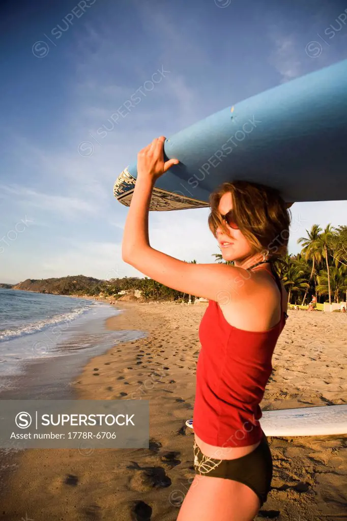 A girl wearing a red top and bikini bottoms holds a surfboard over her head on a remote beach in Sayulita, Mexico.