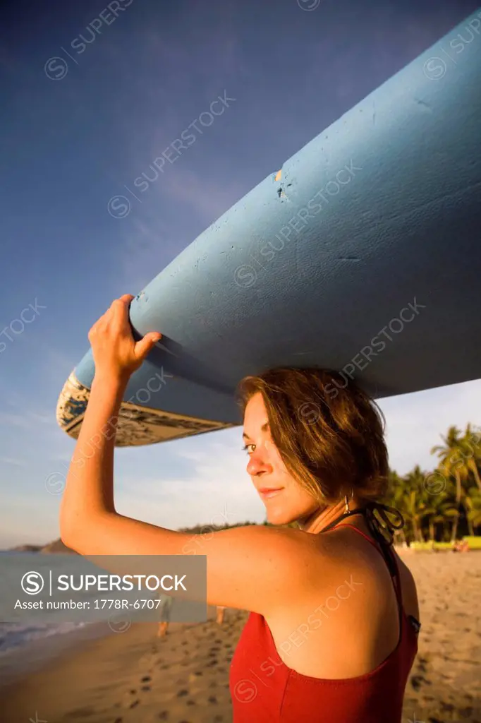 A girl wearing a red top and bikini bottoms holds a surfboard over her head on a remote beach in Sayulita, Mexico.