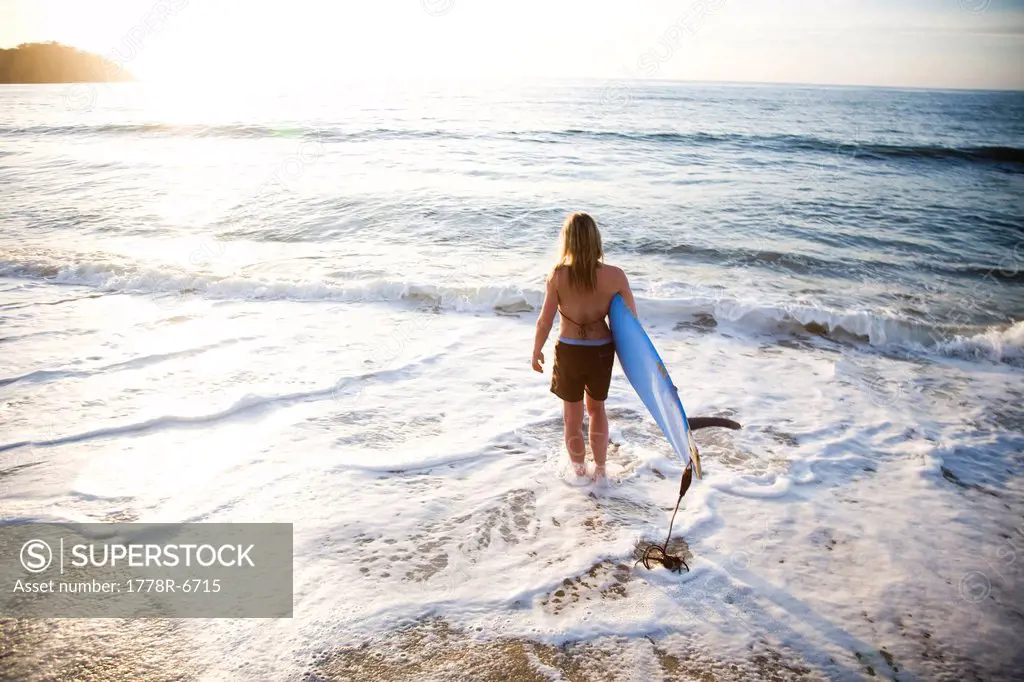 A young woman carries a surfboard into the surf as the sun is setting over the water in Sayulita, Mexico.