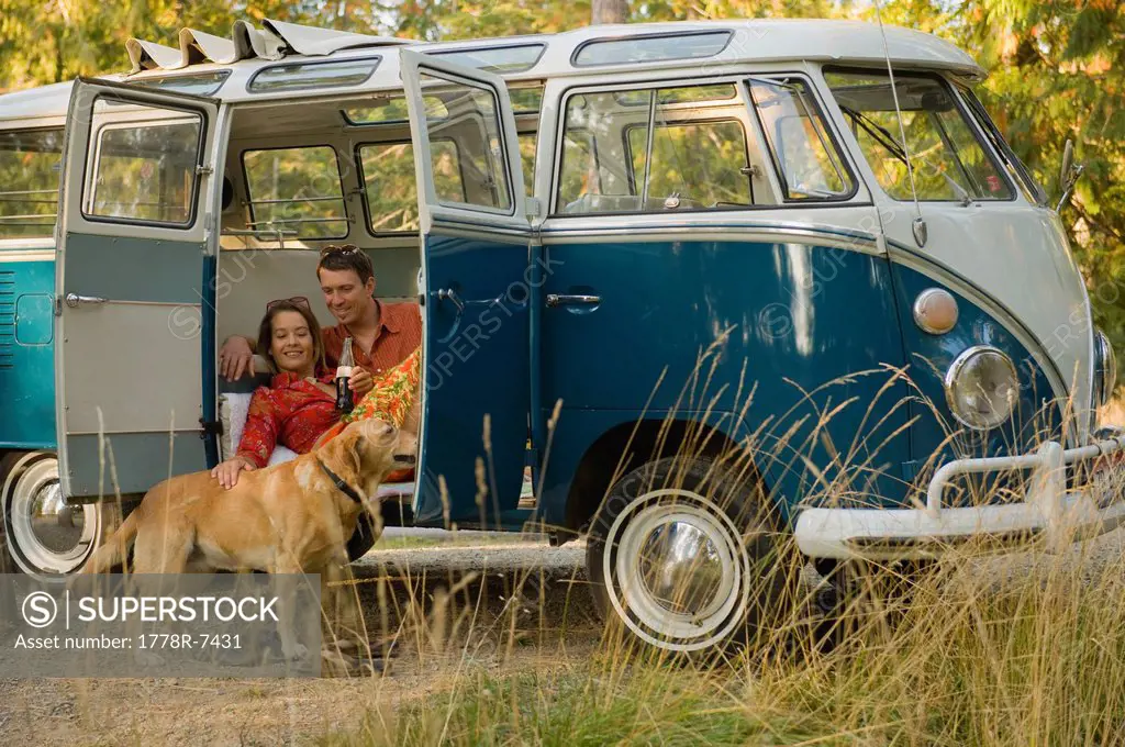 A young couple relax inside a classic van with their dog.