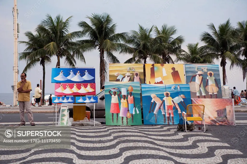 A man selling paintings beside Copacabana beach, with patterned cobblestones on the pavement; Rio de Janeiro, Brazil