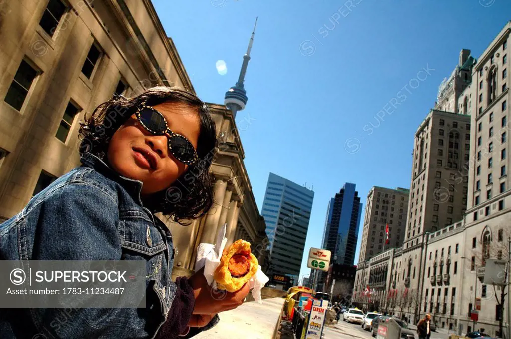 Young girl / child eating hotdog in front of buildings, CN Tower in background, Toronto, Canada