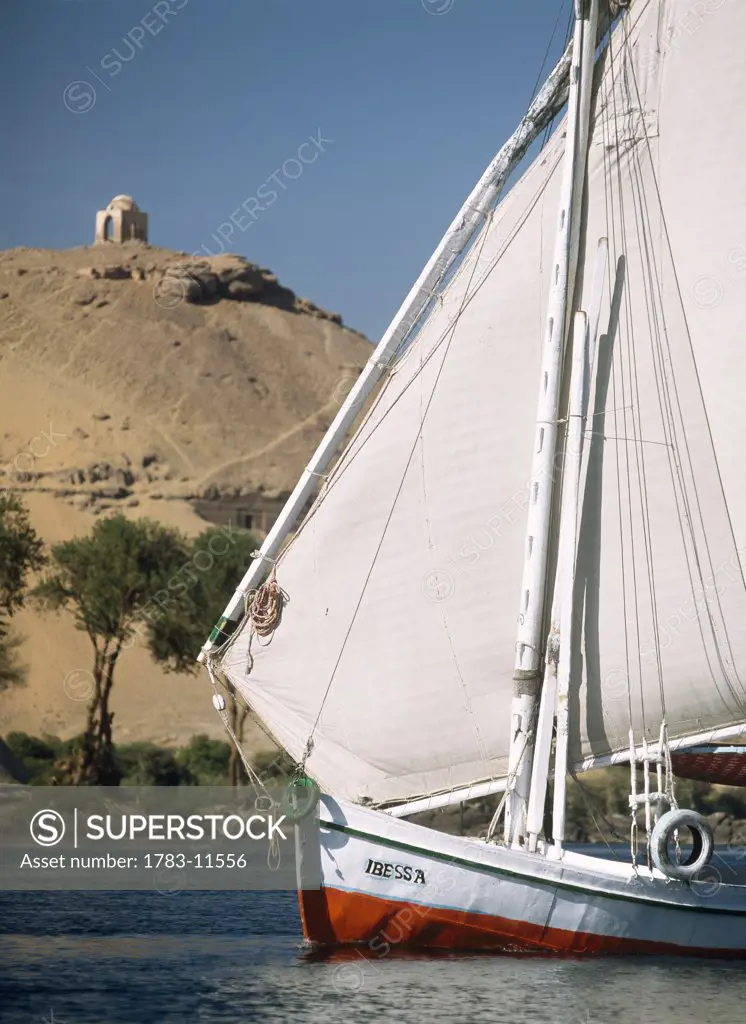 Felucca on River Nile with tombs of nobles and Qubbet el-Hawa behind, Aswan, Egypt.