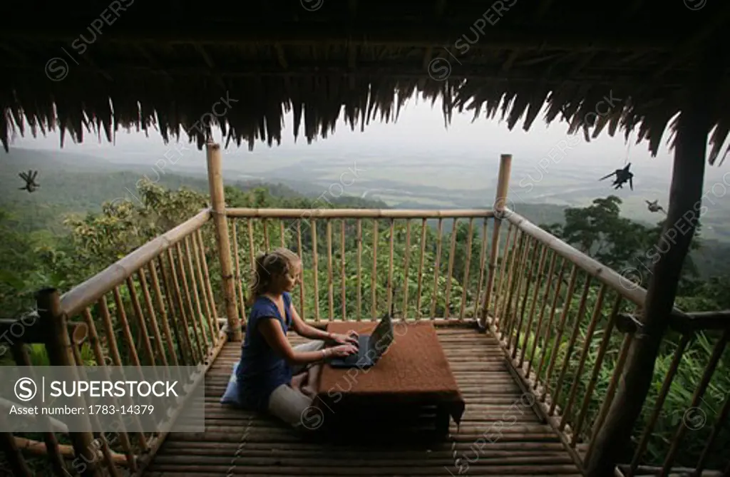 A  woman sitting on the floor using a wireless laptop computer to get email on the balcony of a remote bamboo hut overlooking Bangladesh , Near Mawlynnong , East Khasi Hills , Meghalaya , North East States , India