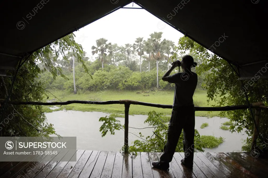 Woman looking through binoculars from tent as the rain comes down in a safari lodge, Liwonde National Park, Malawi