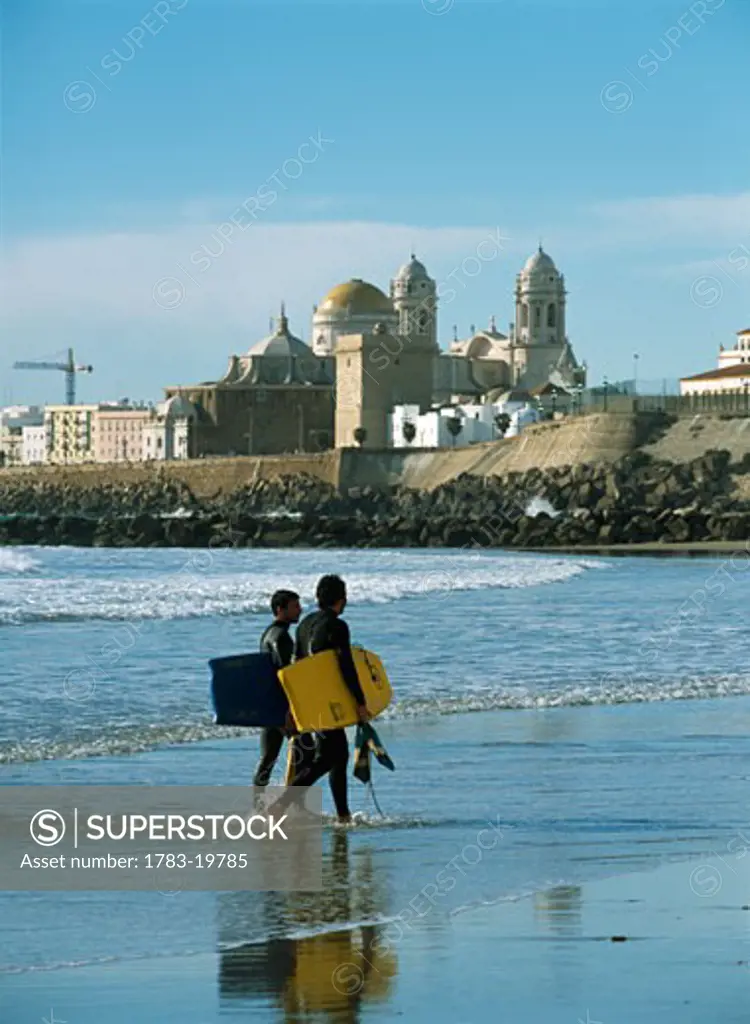 Surfers walking ashore in front of Santa Cruz cathedral, Cadiz, Andalucia, Spain.
