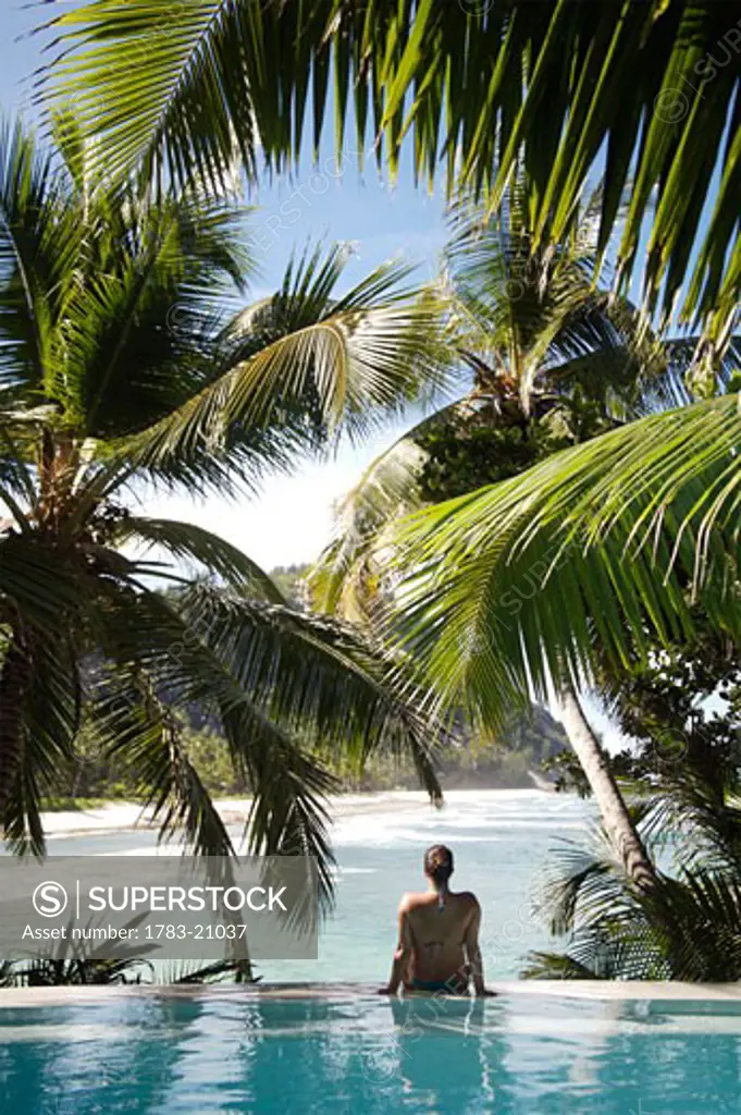 Woman sitting on edge of infinity pool overlooking coastline, North Island, Seychelles