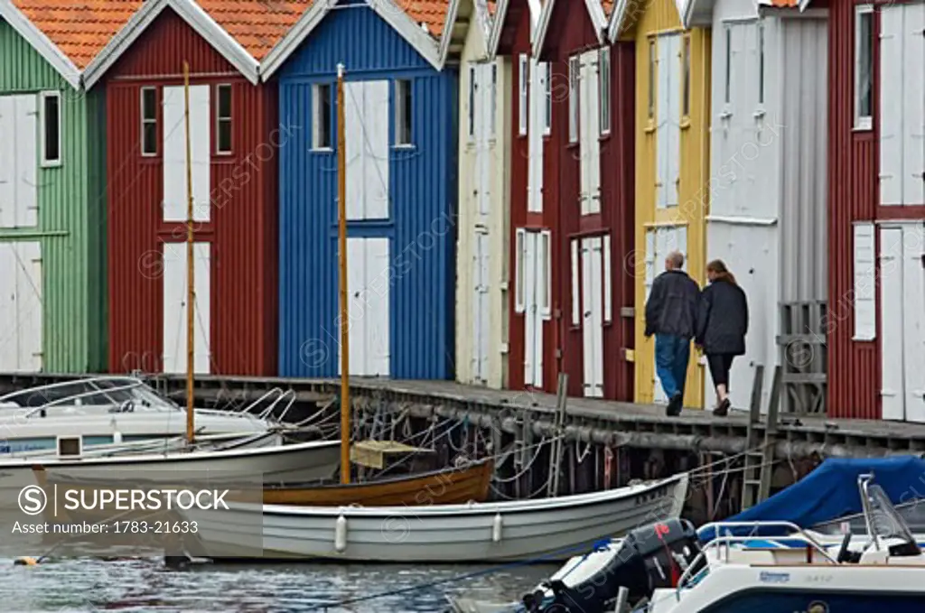 A couple strolling past the colourful fishing huts along the quayside at Smogen. , Sotenas Municipality, Sweden.