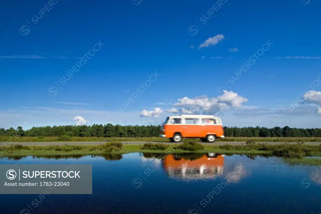 Orange campervan driving past pond, New Forest, Hampshire