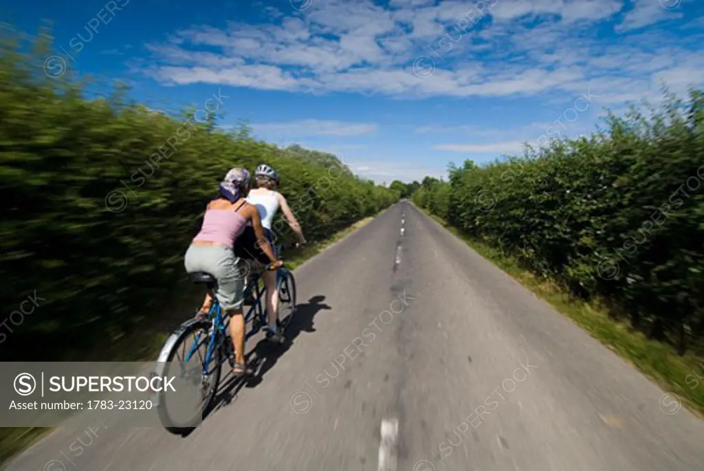 Two women cycling on tandem down road in the countryside, Kent, England