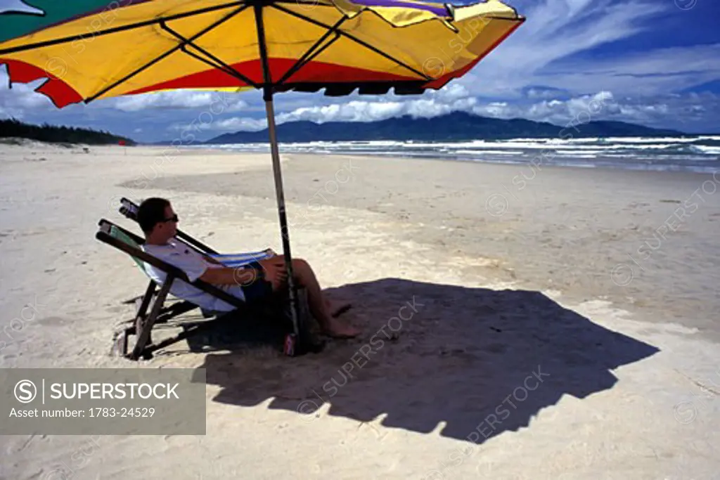 Man sitting on deckchair under umbrella on beach, side view, Non Nuoc Beach, Danang, Vietnam.