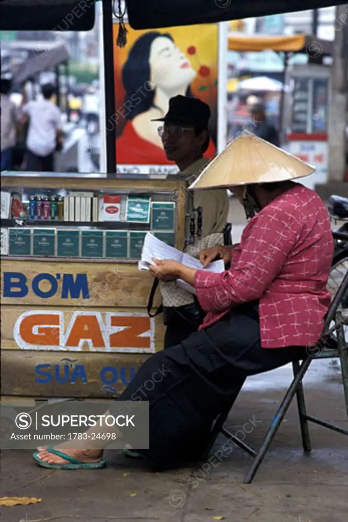Stallholder in conical hat reading newspaper, Ho Chi Minh City, Vietnam.