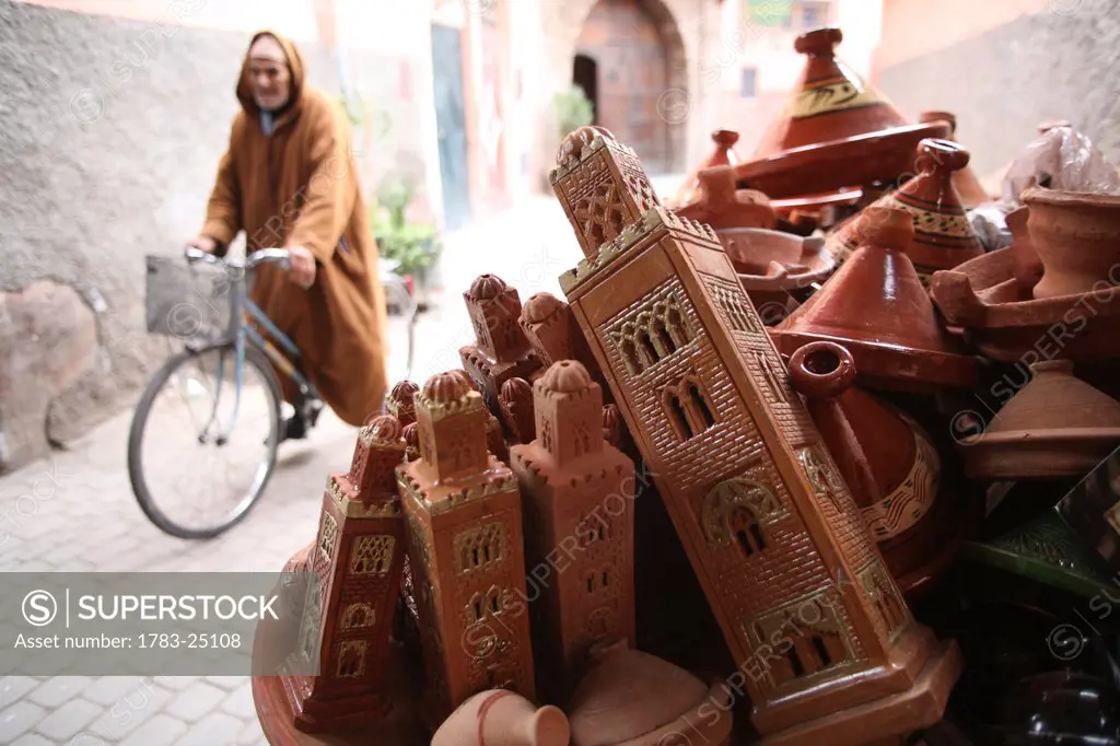 Local man wearing long_hooded cloak, known as a djellaba, passing on his bicycle. Tajine pots and models of minarets of Koutoubia Mosque for sale at a...