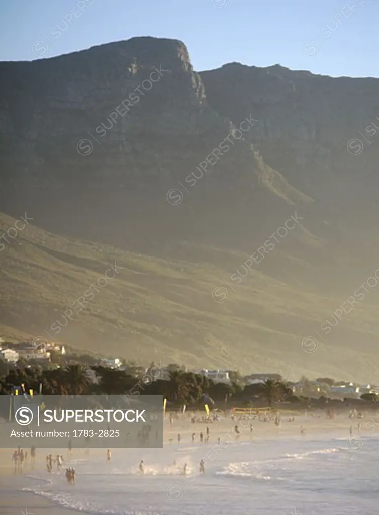 People swimming in the sea in Camps Bay beneath some of The 12 Apostles at dusk, Cape Town, South Africa. 