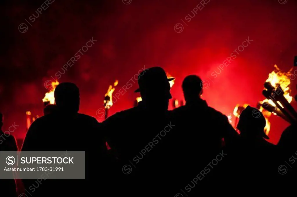 Silhouettes of people in front of red flare and burning torches at Newick Bonfire nightNewick, East Sussex, England. Silhouettes of people in front of red flare and burning torches at Newick Bonfire night