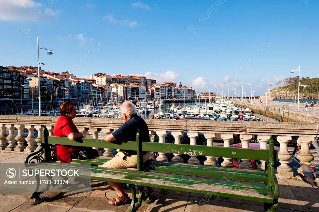 Senior Couple Sitting On Bench At Lekeitio Harbor, Basque Country, Spain