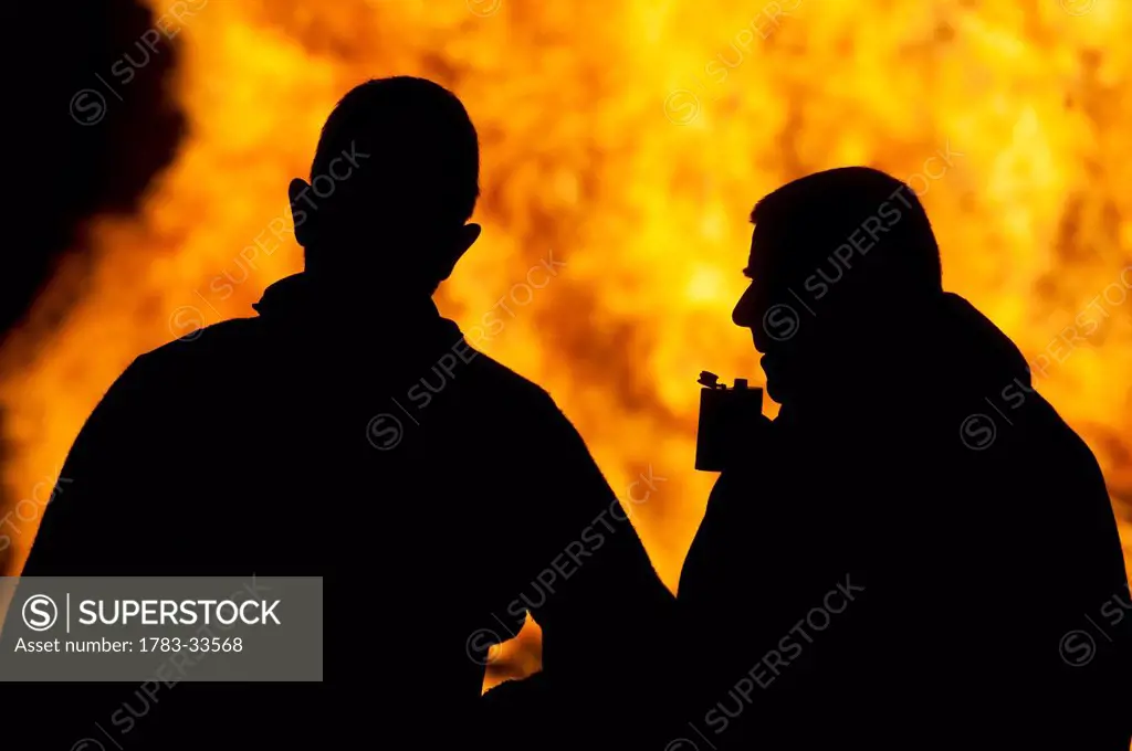 Silhouettes Of Two People, One With Hipflask, In Front Of Large Bonfire At The Ewhurst And Staplecross Bonfire Night, East Sussex, Uk