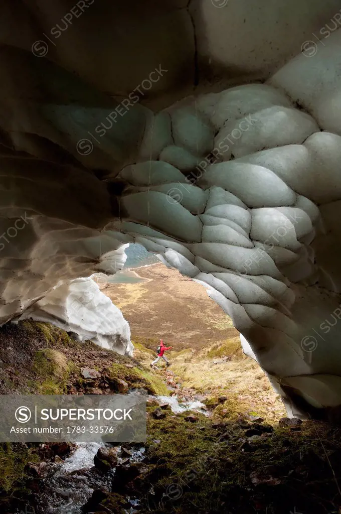 Walker Jumping Over Small Stream Coming Out Of Ice Cave, Gleann Einich Near Aviemore, Highlands, Scotland, Uk